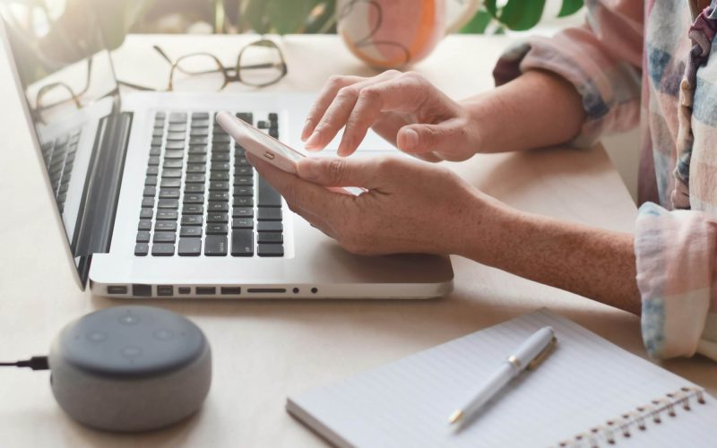 woman-sitting-at-desk-with-laptop-computer-and-virtual-assistant-smart-speaker-using-mobile-phone.jpg
